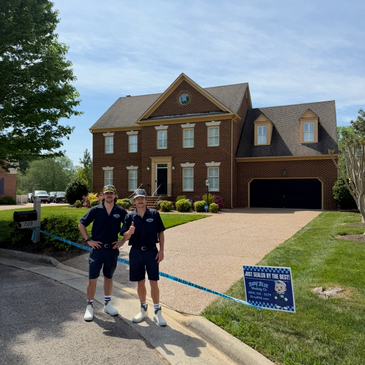 Two men in navy uniforms stand in front of a large brick house with a manicured lawn.