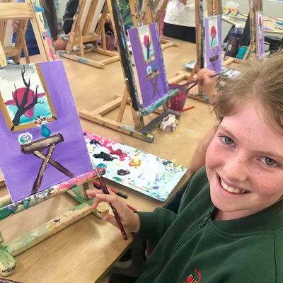 Smiling girl painting a vibrant landscape with a sunset and tree indoors.