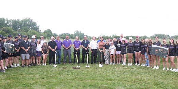 A large group of people posing outdoors with shovels on a grassy field.