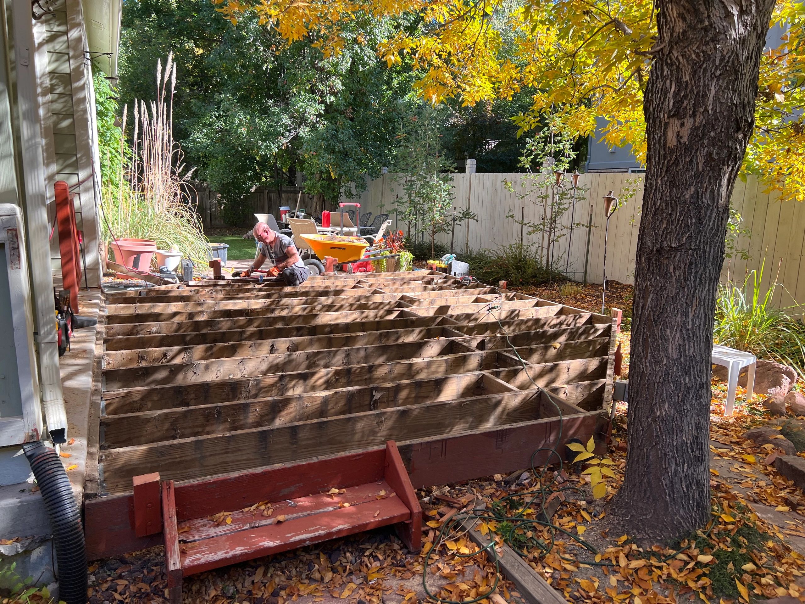 Man building a wooden deck in a sunny backyard with autumn leaves.