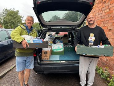 Two men holding boxes of supplies standing by an open car trunk.