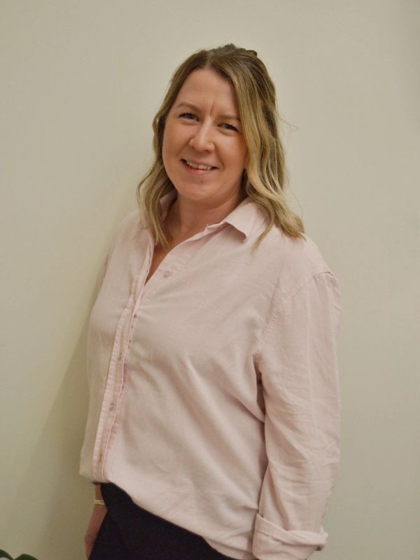Smiling woman in a light pink shirt standing against a plain wall.