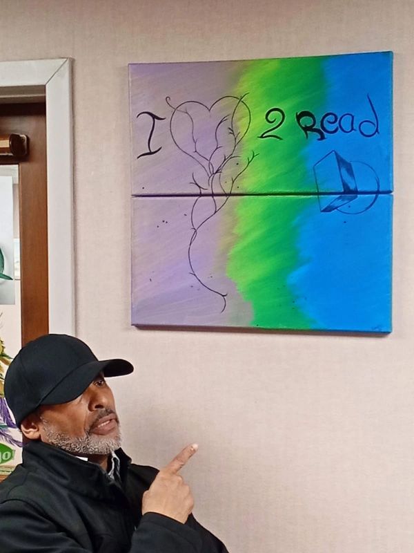Man points to colorful painting saying 'I love 2 read' with a heart and book.