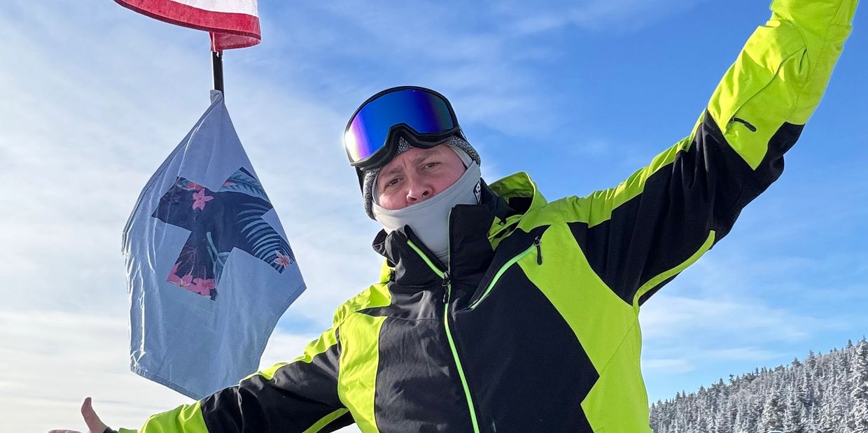 Person in bright ski jacket posing excitedly on a snowy mountain with flags in the background.