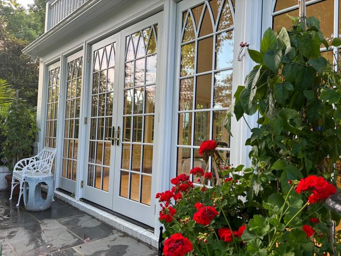 White French doors open to a patio with red flowers and a white bench.