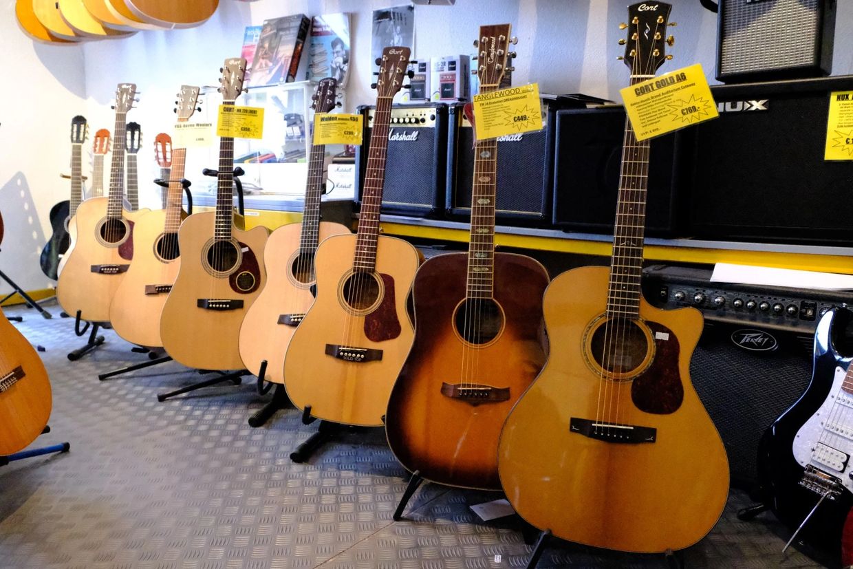 Akustikgitarren, die nebeneinander auf Gitarrenständer gestellt sind. Dahinter stehen Verstärker.