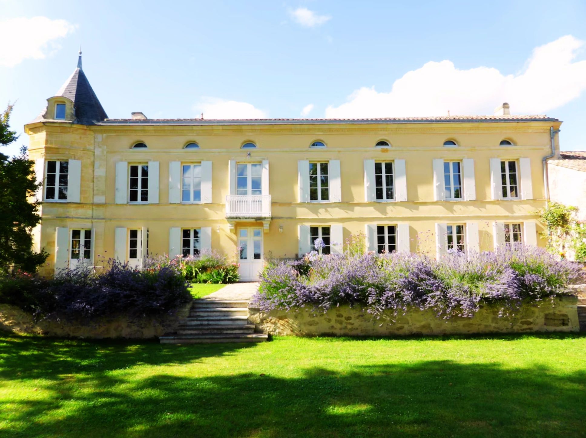 Elegant yellow mansion with white shutters and purple flowers in front.