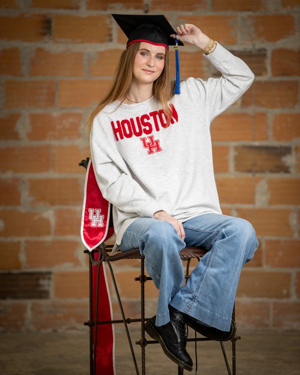 Graduate in a Houston sweatshirt and cap, sitting confidently on a rustic chair.