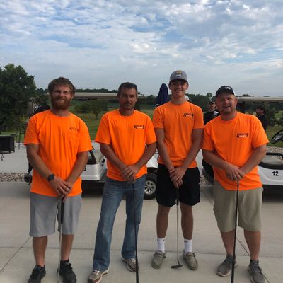 Four men in bright orange shirts holding golf clubs posing outdoors by golf carts.