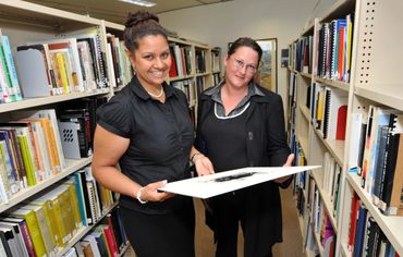 Two women examining a large sheet in a library aisle.