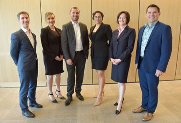 Six professionals posing in business attire against a wooden wall.