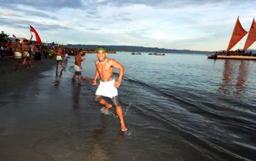 Man in traditional attire running on the beach during a cultural event with boats in the background.