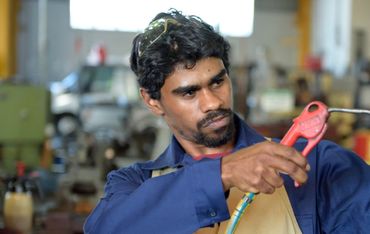 A man in a workshop sprays air on a metal part.