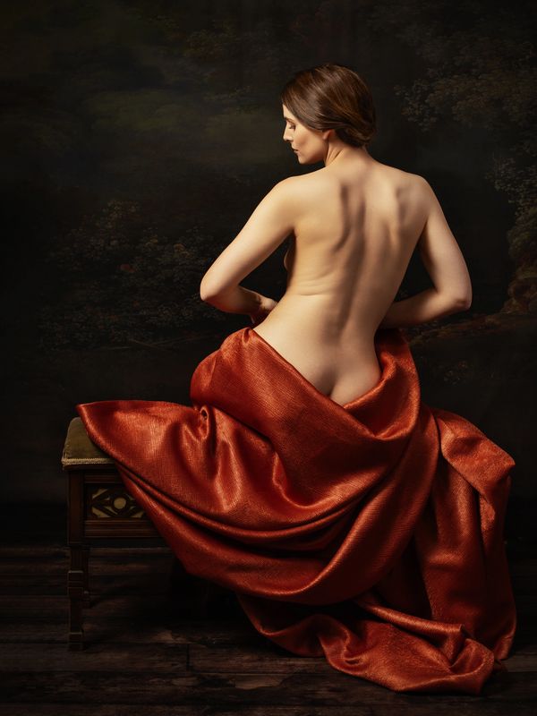 A woman with bare back draped in rich red fabric sits on a wooden bench.