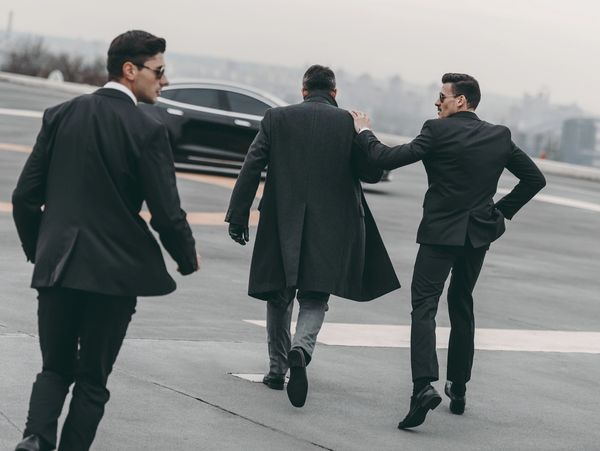 Three men in suits walking on a helipad with a black car in the background.
