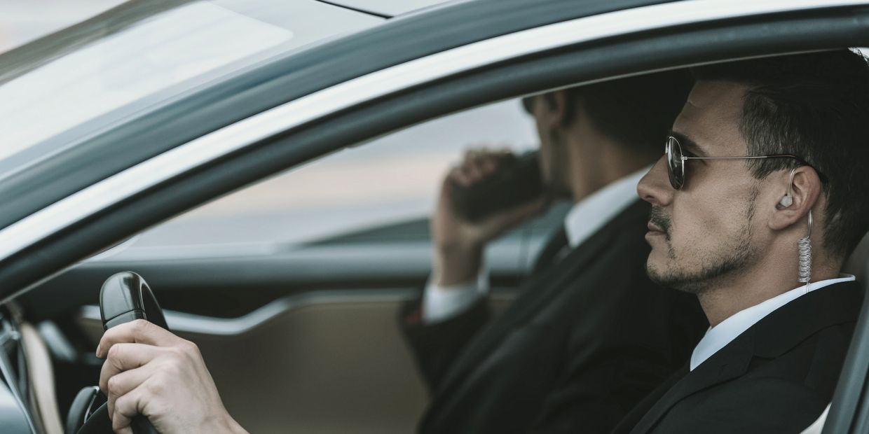 Two men in suits with earpieces inside a car, one driving.