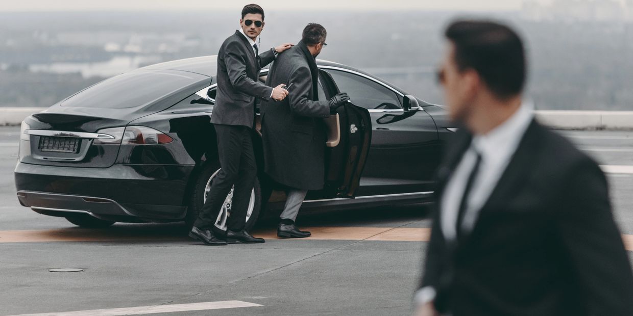 Two men in black suits with sunglasses beside a black car, one opening the door for the other.