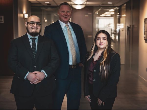 Three professionally dressed individuals posing in a modern office hallway.