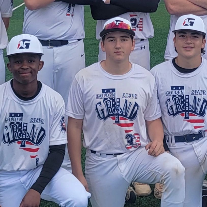Three young baseball players kneeling and smiling, wearing matching uniforms.