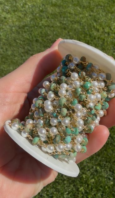 Hand holding a spool of decorative beaded chain with pearls and green stones.