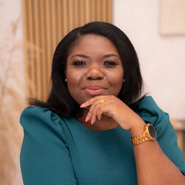 A smiling Black woman wearing a teal blouse sits confidently at a desk with her hand resting on her 