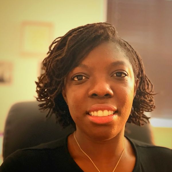 A smiling Black woman with short hair wears a dark top and looks directly at the camera in a well-li