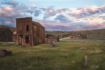 Old wooden buildings in a quiet, grassy ghost town at sunset.