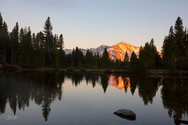 Sunset illuminates mountain peak reflected in calm lake surrounded by trees.