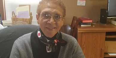 An elderly woman with glasses smiles warmly in an office setting.