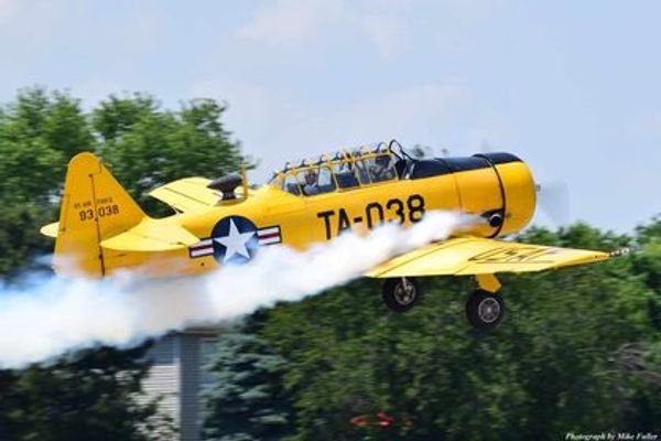 Yellow vintage military aircraft flying with smoke trail.
