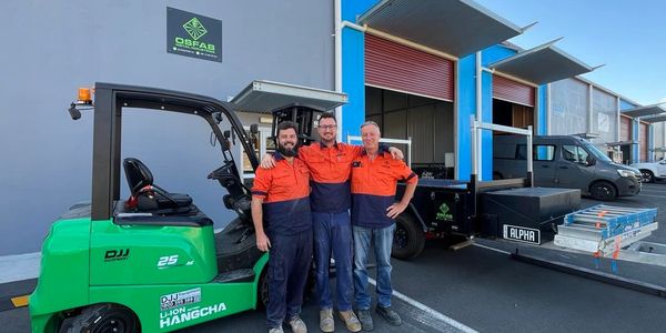 Three workers in orange uniforms stand smiling by a green forklift outside a warehouse.