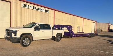 White pickup truck towing a long flatbed trailer in an industrial area.