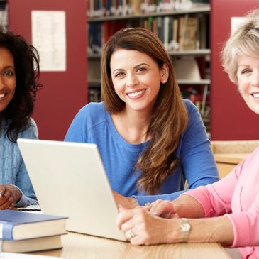 Three women smiling around a laptop in a library setting.