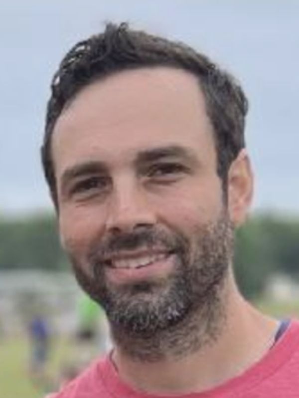 Smiling man with dark hair and beard outdoors.