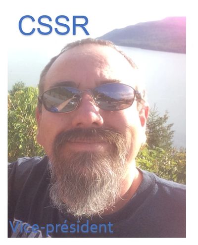 Man with sunglasses and beard smiling outdoors near water.