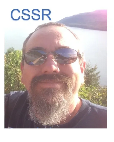 Man with sunglasses and beard smiling outdoors near water and trees.