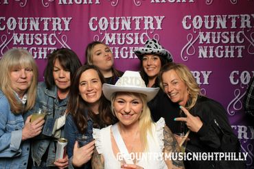 A group of women smiling at a country music night event with a bride in a white hat.