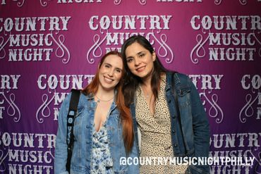 Two women smiling in front of a Country Music Night backdrop.