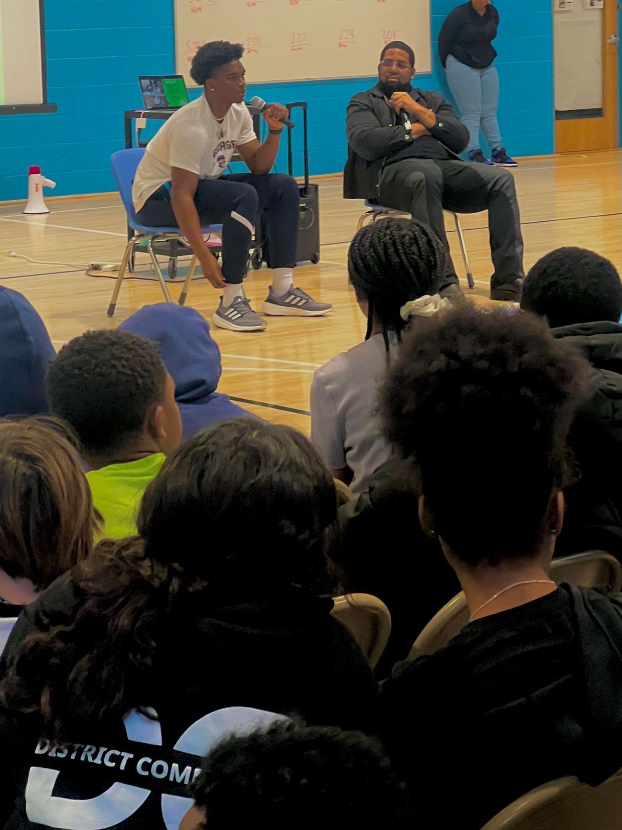Two men speaking to an audience in a gymnasium.