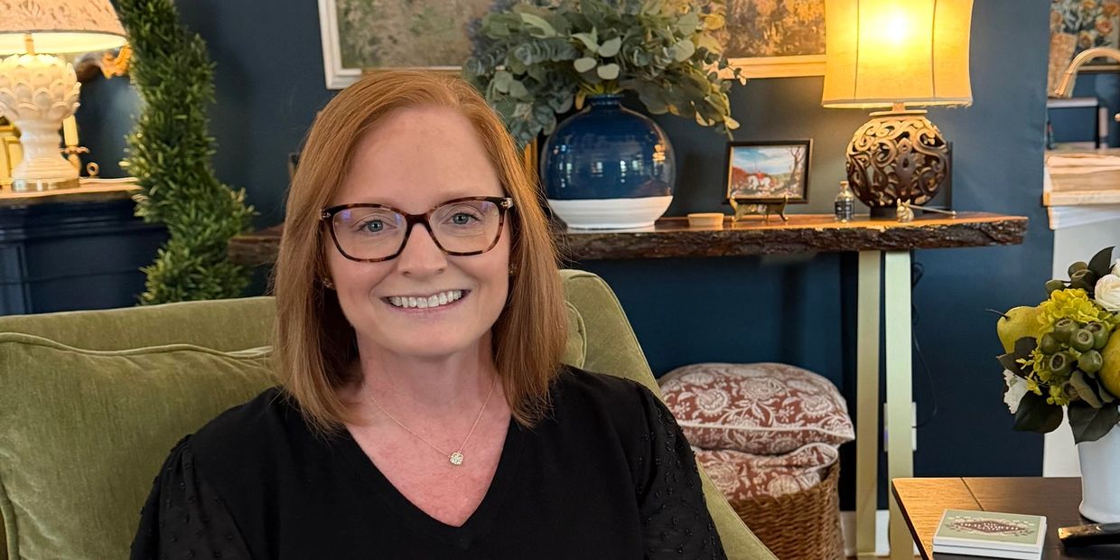 Smiling woman with red hair and glasses sitting on a green couch in a cozy, warmly lit room.