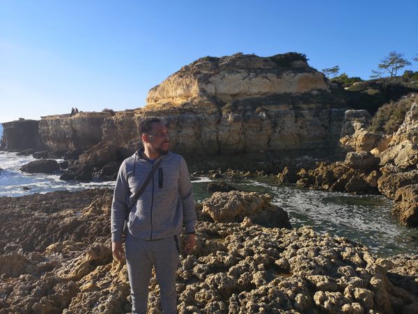 A man in gray sportswear stands on rocky terrain near the ocean under a clear blue sky.
