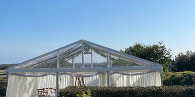 A clear tent with white curtains set in a garden under a clear blue sky.