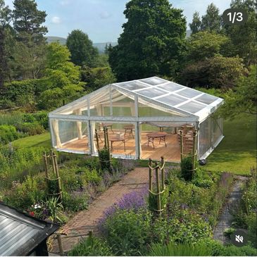Transparent tent set up in a lush garden with wooden furniture inside.