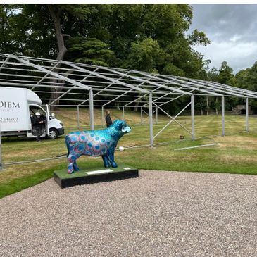 Colorful cow sculpture stands near metal framework outdoors.