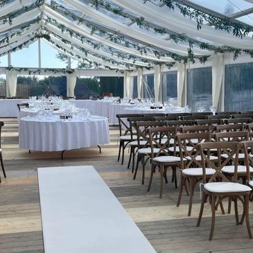 Elegant wedding reception setup inside a clear tent with white decorations and wooden chairs.