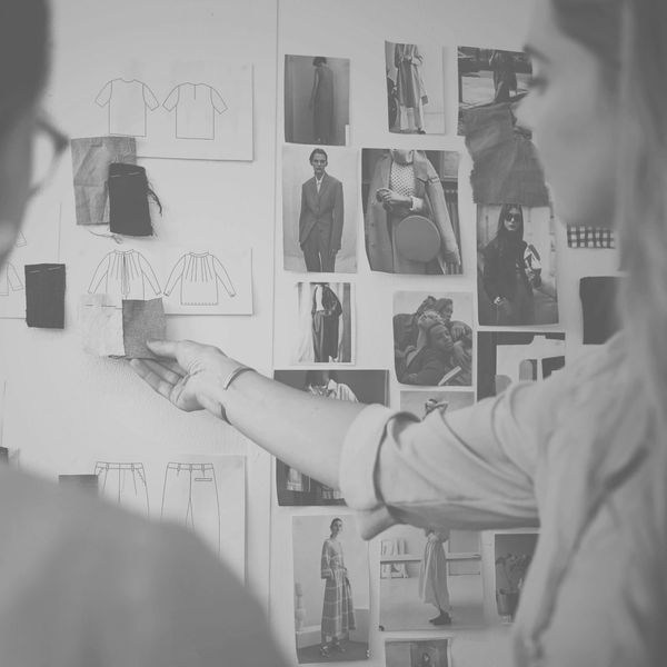 Two women review fabric swatches and fashion sketches on a wall.