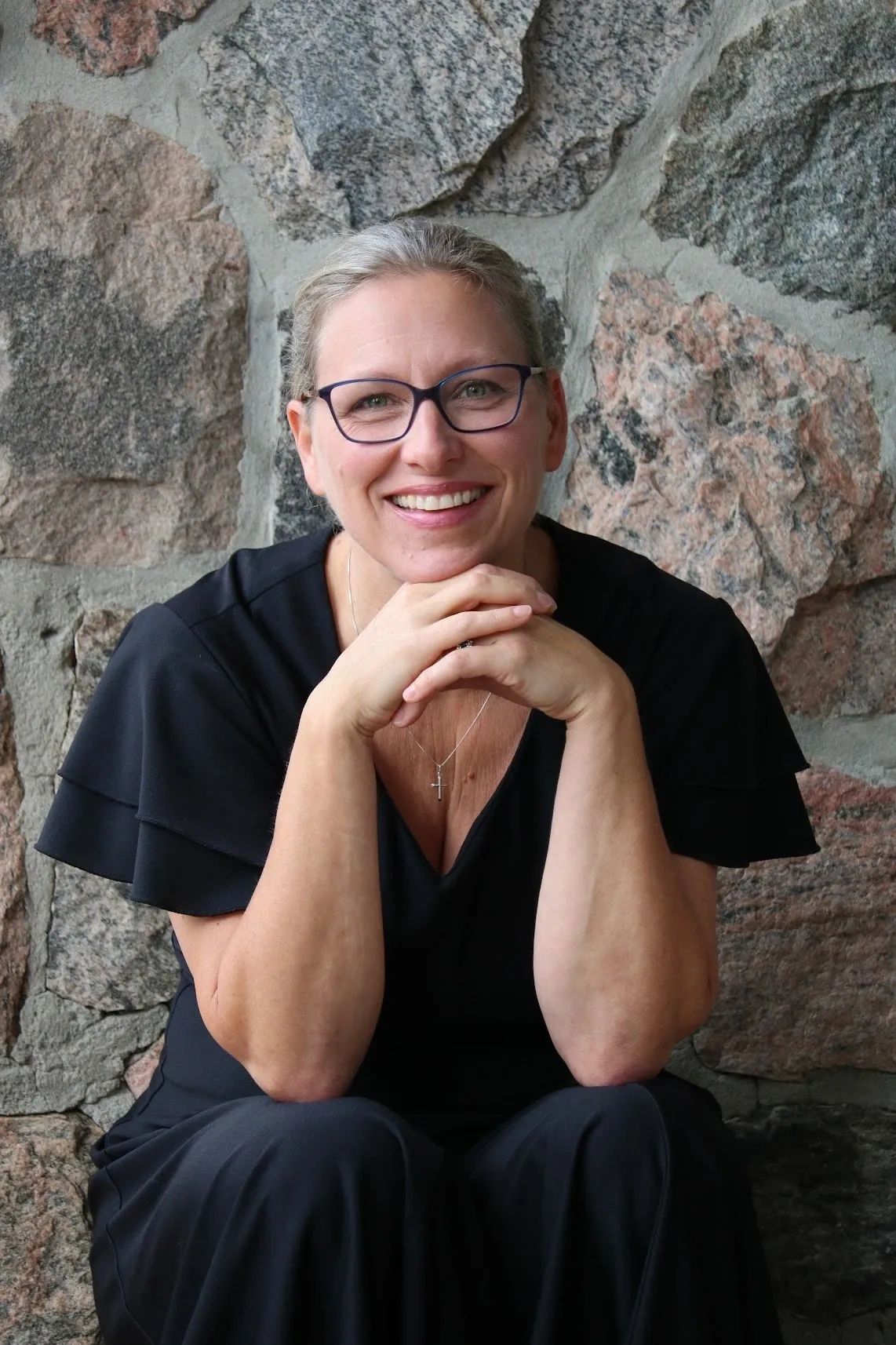 Smiling woman in glasses sitting against a stone wall, wearing black.