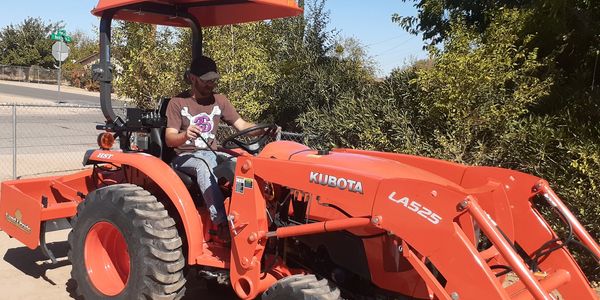 Person operating an orange Kubota tractor outdoors on a sunny day.