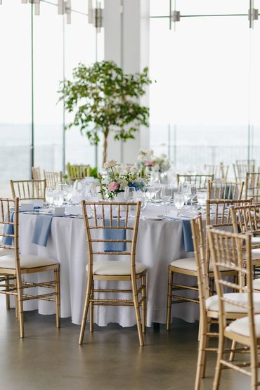 Round table, flanked with gold chiavari chairs, set with white table cloths and light blue napkins. 
