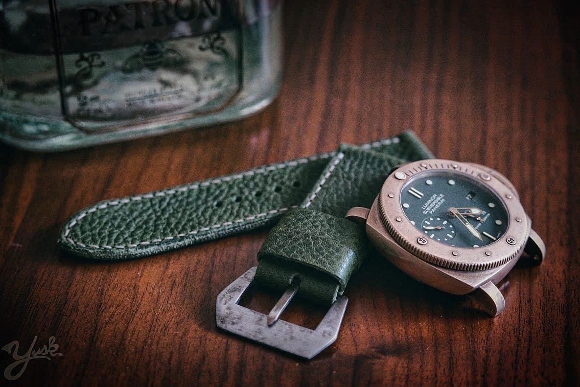Close-up of a green leather watch strap with a bronze watch face on a wooden surface.